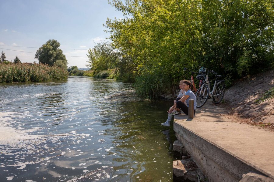 Zwei Frauen sitzen direkt am Fluss in der Sonne, die Fahrräder stehen abgestellt im Hintergrund