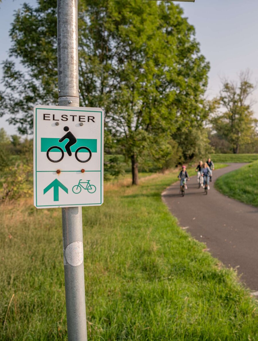 Radwegeschild zum elsterradweg, im Hintergrund fährt eine Familie auf dem Radweg entlang