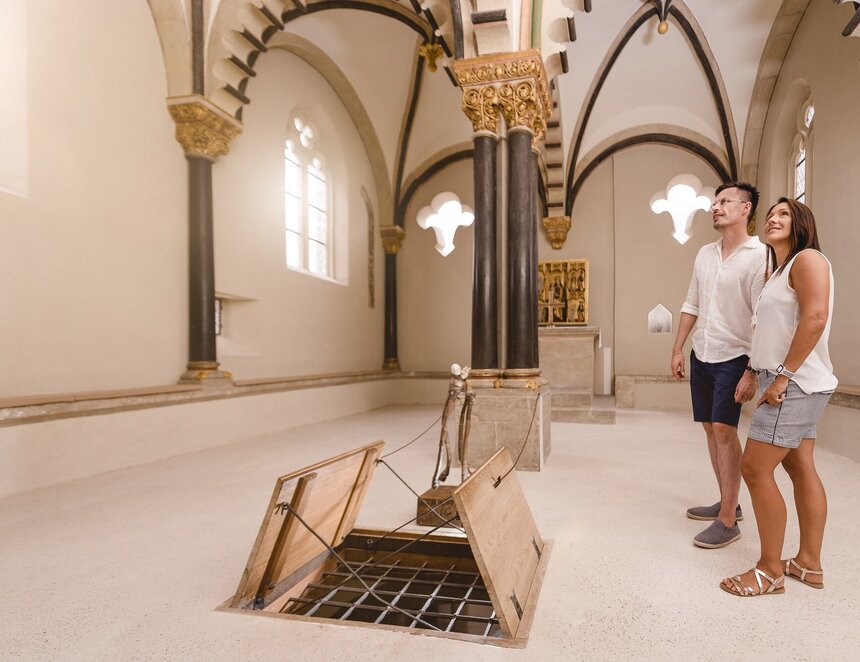 Ein Paar besucht die Doppelkapelle Schloss Neuenburg. Es ist ein weißer Raum mit romanischen Fenstern und einer geöffneten Bodenklappe.